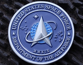 May include: A blue and silver United States Space Force coin. The coin features the text "United States Space Force" and "Department of the Air Force" around the edge. The center design includes a stylized delta and a globe.