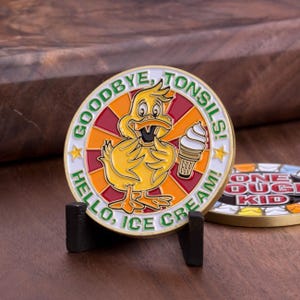 Pode incluir: Uma moeda dourada com um pato amarelo segurando um cone de sorvete. A moeda tem o texto "Goodbye, Tonsils! Hello, Ice Cream!" ao redor da borda.