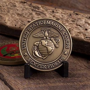 Puede incluir: Una moneda conmemorativa del Cuerpo de Marines de los Estados Unidos de color bronce. La moneda presenta un águila, un globo terráqueo y un ancla, con las palabras "United States Marine Corps" y "Semper Fidelis". La moneda se exhibe en un pequeño soporte negro.