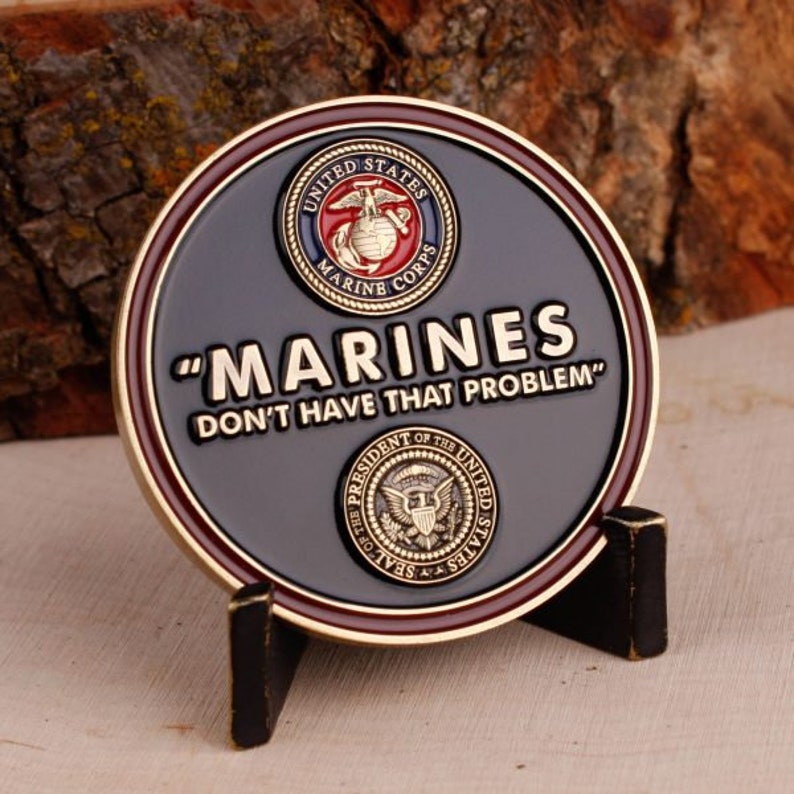 May include: A round, dark gray coin with gold and burgundy trim, featuring the words "MARINES DON'T HAVE THAT PROBLEM." The coin displays the United States Marine Corps emblem and the Presidential Seal.
