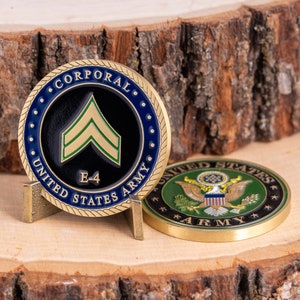 以下が含まれることがあります： 金色のコインで、青と黒のデザインで、「Corporal、United States Army、E-4」とシェブロンの記章が書かれています。コインの裏面は緑と金のデザインで、「United States Army」とハクトウワシが書かれています。