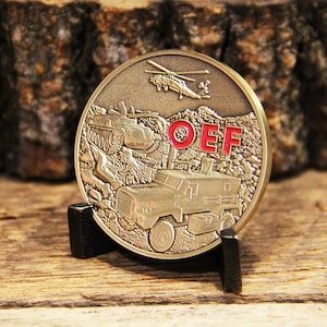 May include: A gold-colored challenge coin with a helicopter, two military vehicles, and the letters "OEF" in red enamel. The coin is on a black stand.