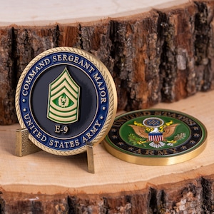 Puede incluir: Dos monedas conmemorativas, una de pie en un soporte, la otra plana. La primera moneda presenta el texto "COMMAND SERGEANT MAJOR" y "UNITED STATES ARMY" alrededor de un emblema central. La segunda moneda muestra el emblema del Ejército de EE. UU.