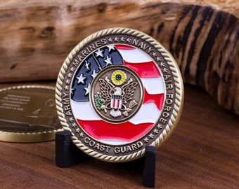 May include: A gold-colored commemorative coin featuring the emblems of the U.S. Army, Marines, Navy, Air Force, and Coast Guard. The coin has a red, white, and blue design with a central eagle emblem. It is displayed on a small black stand.