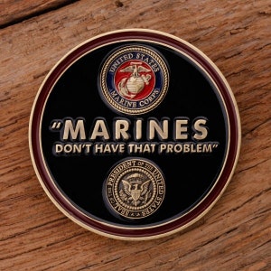 May include: A round, black coin with a gold rim and a maroon border. The coin features the words "MARINES DON'T HAVE THAT PROBLEM" in gold lettering. The United States Marine Corps emblem and the Presidential Seal are also displayed.