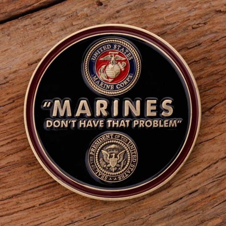 May include: A round, black coin with a gold rim and a maroon border. The coin features the words "MARINES DON'T HAVE THAT PROBLEM" in gold lettering. The United States Marine Corps emblem and the Presidential Seal are also displayed.
