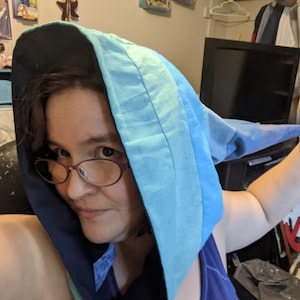 May include: A person wearing a blue and black hooded garment with a long tail. The hood is pulled up over their head, and they are looking at the camera.