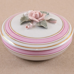 Pode incluir: Uma caixa de porcelana branca para bijuterias com uma rosa rosa e folhas na tampa. A caixa tem listras rosa e douradas ao redor da borda.