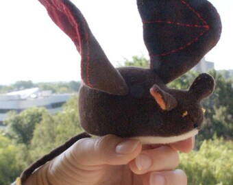 May include: A brown plush toy with a long tail and wings. The wings are dark brown with red stitching. The toy is being held in a hand.