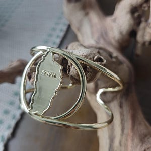 May include: A gold-colored cuff bracelet featuring a cutout of a geographical shape, possibly a state or region, within a circular frame. The word "KOREA" is visible within the cutout. The bracelet has a simple, elegant design.