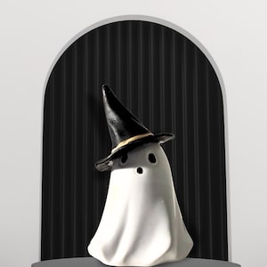 May include: A white ghost figurine wearing a black witch hat with a thin gold band around the base. The ghost has two black oval eyes and a small black circular mouth, with a smooth, draped body shape that flares out at the bottom edge.