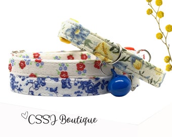 May include: Two floral patterned cat collars with a blue bell charm. The collars are white with red and blue flowers. The top collar has yellow flowers. The collars are stacked on top of each other. The text "CSSJ Boutique" is visible in the bottom right corner of the image.