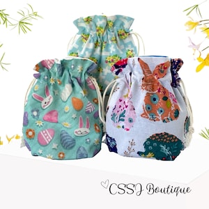 May include: Three drawstring bags with different Easter-themed patterns. One bag has a blue background with white stars and crosses, another has a green background with pink eggs and bunnies, and the third has a white background with colorful bunnies and flowers. The bags are made of fabric and have a drawstring closure.