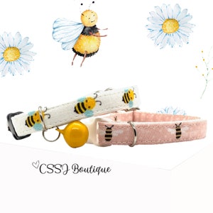 May include: Two cat collars with a bee pattern. One collar is white with blue bees and the other is pink with black bees. Both collars have a bell.
