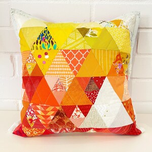 Könnte beinhalten: Ein quadratisches Kissen mit einem Patchwork-Muster aus gelben, orangen und roten Dreiecken. Das Kissen ist aus verschiedenen Stoffen mit verschiedenen Mustern gefertigt.