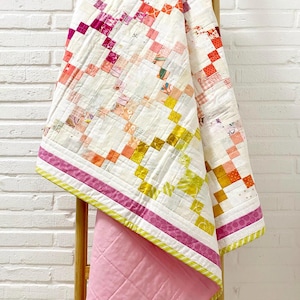 Può includere: Una trapunta fatta a mano con un design patchwork nei toni del rosa, giallo e arancione. La trapunta è drappeggiata su una scala di legno, mostrando il suo intricato motivo e la sua texture trapuntata. Il retro è in tessuto rosa tinta unita.