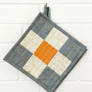 Könnte beinhalten: Ein quadratischer, gesteppter Topflappen mit geometrischem Design. Der Topflappen hat ein zentrales orangefarbenes Quadrat, das von weißen Quadraten mit goldenen Wirbeln und grauen Quadraten mit goldenen Flecken umgeben ist. Eine graue Stoffeinfassung rahmt das Design ein. Der Topflappen hängt an einer weißen Backsteinmauer.