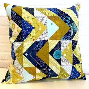 Op de afbeelding: Een vierkant kussen met een geometrisch patroon van blauwe, gele en witte driehoeken. Het kussen is gemaakt van stof met verschillende patronen, waaronder stippen, strepen en bloemenpatronen.