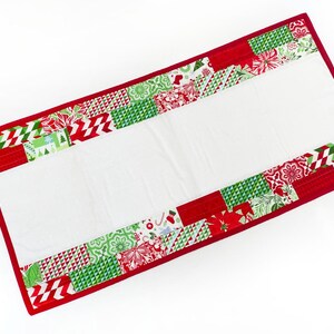 Könnte beinhalten: Rechteckiger Weihnachtstischläufer mit weißer Mitte und roten Rändern. Die Ränder weisen ein Patchwork-Design mit roten, grünen und weißen Mustern auf, darunter geometrische Formen und florale Motive. Der Läufer ist etwa 76 cm lang.