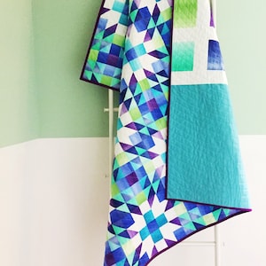 Könnte beinhalten: Eine geometrisch gemusterte Steppdecke in Weiß, Blau, Grün und Lila mit türkisfarbenem Rücken. Die Steppdecke ist gefaltet und hängt an einer weißen Leiter.