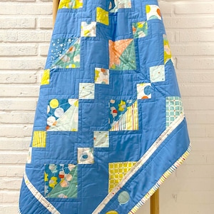 Op de afbeelding: Een handgemaakte babyquilt met een patchwork-ontwerp. De quilt heeft een lichtblauwe achtergrond met vierkanten in verschillende patronen en kleuren, waaronder geel, wit en oranje. De rand is effen lichtblauw met een gestreepte rand en ballonapplicaties.