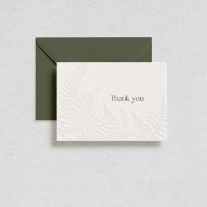 May include: A white rectangular greeting card with an embossed fern leaf pattern and the text Thank you in dark gray ink, paired with a solid dark olive green envelope. The card features a textured surface with raised botanical details.