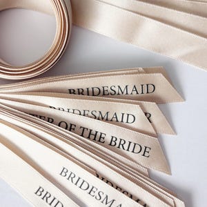 Puede incluir: Un carrete de cinta de satén color champán con varios extremos sueltos que se extienden hacia afuera. Cada extremo de la cinta presenta texto impreso en negro que incluye las palabras BRIDESMAID y MOTHER OF THE BRIDE, con un corte en ángulo.