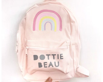 May include: A pink canvas backpack with a rainbow design and the name "DOTTIE BEAU" printed on the front.