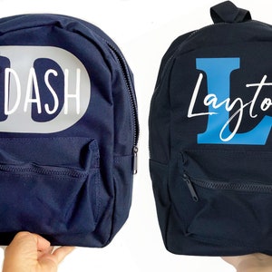May include: Two navy blue canvas backpacks with personalized names. The first backpack has a large white letter D with the name "DASH" written in white. The second backpack has a large blue letter L with the name "Layton" written in white.