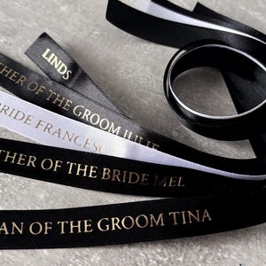 May include: A collection of black and white satin ribbons featuring gold foil text. The ribbons are printed with titles including MOTHER OF THE GROOM JULIE, BRIDE FRANCESCA, MOTHER OF THE BRIDE MEL, NAN OF THE GROOM TINA, and LINDSEY. The ribbons are smooth.