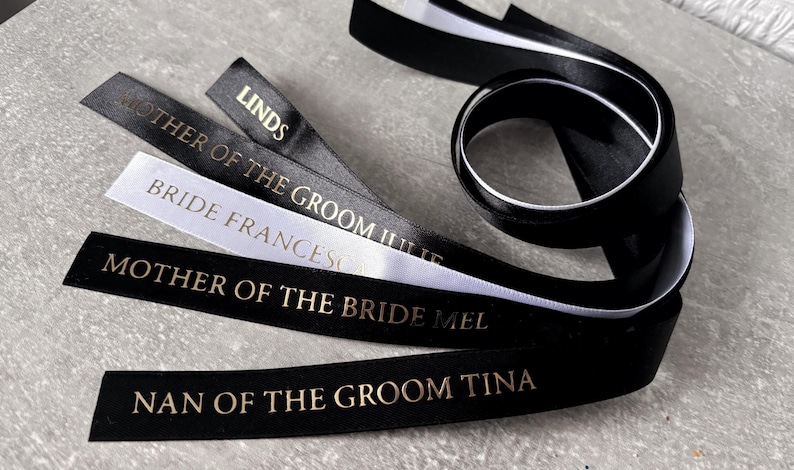 May include: A collection of black and white satin ribbons featuring gold foil text. The ribbons are printed with titles including MOTHER OF THE GROOM JULIE, BRIDE FRANCESCA, MOTHER OF THE BRIDE MEL, NAN OF THE GROOM TINA, and LINDSEY. The ribbons are smooth.