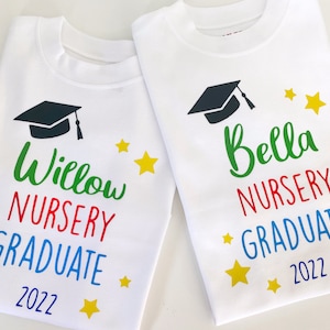 May include: Two white t-shirts featuring a black graduation cap graphic, yellow stars, and colourful text. One shirt reads Willow NURSERY GRADUATE 2022, and the other reads Bella NURSERY GRADUATE 2022, with names in green, nursery in red, and graduate in blue.