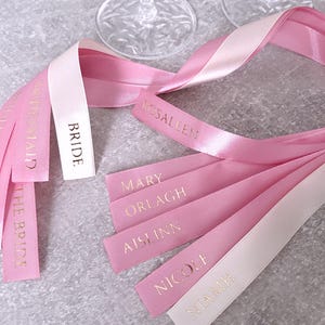 May include: A collection of satin ribbons in shades of light pink and white, featuring gold foil text. The ribbons are printed with the words BRIDE, BRIDESMAID, ROSALEEN, MARY, ORLAGH, AISLINN, NICOLE, and NIAMH, arranged in a loose, flowing cluster.