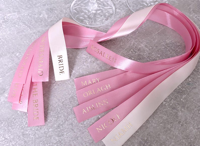 May include: A collection of satin ribbons in shades of light pink and white, featuring gold foil text. The ribbons are printed with the words BRIDE, BRIDESMAID, ROSALEEN, MARY, ORLAGH, AISLINN, NICOLE, and NIAMH, arranged in a loose, flowing cluster.