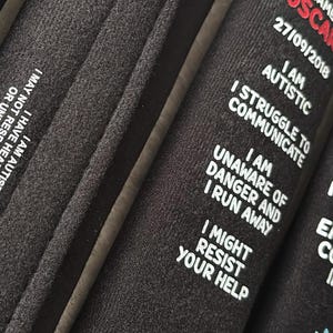 May include: Three black fabric wristbands featuring white and red printed text. The text includes personal information and safety warnings such as I AM AUTISTIC, I STRUGGLE TO COMMUNICATE, I AM UNAWARE OF DANGER AND I RUN AWAY, and EMERGENCY CONTACTS INSIDE.