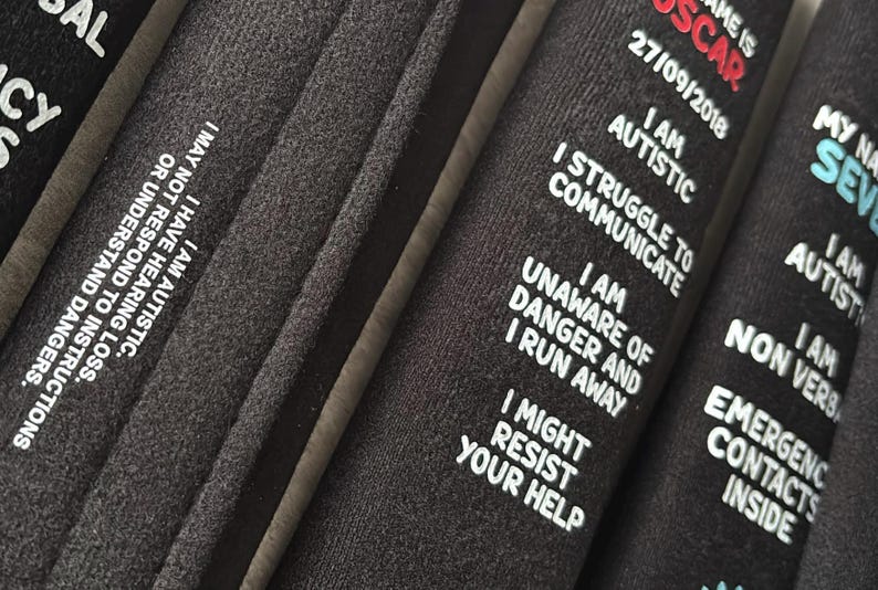 May include: Three black fabric wristbands featuring white and red printed text. The text includes personal information and safety warnings such as I AM AUTISTIC, I STRUGGLE TO COMMUNICATE, I AM UNAWARE OF DANGER AND I RUN AWAY, and EMERGENCY CONTACTS INSIDE.