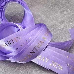 May include: A lavender satin ribbon featuring gold foil text that reads SAMANTHA & MARTIN and MAY 2026. The ribbon is coiled in a loose loop, displaying a smooth, reflective texture and a repeating printed pattern along its length.