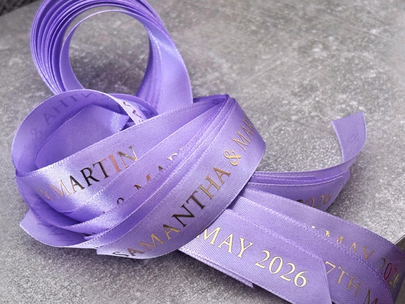 May include: A lavender satin ribbon featuring gold foil text that reads SAMANTHA & MARTIN and MAY 2026. The ribbon is coiled in a loose loop, displaying a smooth, reflective texture and a repeating printed pattern along its length.