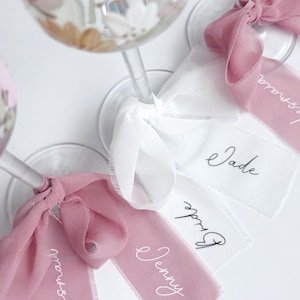May include: Clear glass stems featuring hand-painted floral designs in muted tones. Each glass is adorned with a tied fabric ribbon in dusty rose or white, featuring printed names and titles including Jade, Jenny, Bride, and Bridesmaid.