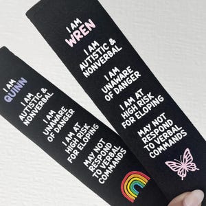 May include: Two black fabric seatbelt covers featuring white text that reads: I AM QUINN or I AM WREN, I AM AUTISTIC & NONVERBAL, I AM UNAWARE OF DANGER, I AM AT HIGH RISK FOR ELOPING, MAY NOT RESPOND TO VERBAL COMMANDS. One has a rainbow, one a butterfly.