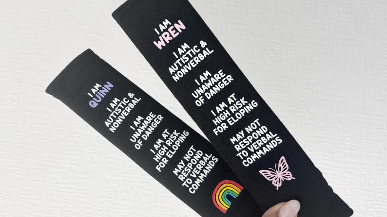 May include: Two black fabric seatbelt covers featuring white text that reads: I AM QUINN or I AM WREN, I AM AUTISTIC & NONVERBAL, I AM UNAWARE OF DANGER, I AM AT HIGH RISK FOR ELOPING, MAY NOT RESPOND TO VERBAL COMMANDS. One has a rainbow, one a butterfly.