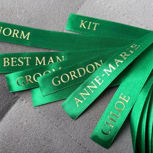 May include: A collection of emerald green satin ribbons featuring gold foil lettering. The ribbons are printed with the names NORM, KIT, GORDON, ANNE-MARIE, CHLOE, and the titles BEST MAN and GROOM, arranged in a layered, overlapping fan pattern.