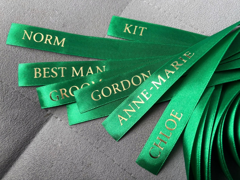 May include: A collection of emerald green satin ribbons featuring gold foil lettering. The ribbons are printed with the names NORM, KIT, GORDON, ANNE-MARIE, CHLOE, and the titles BEST MAN and GROOM, arranged in a layered, overlapping fan pattern.