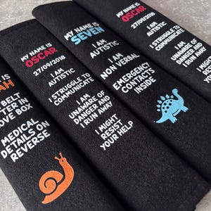 May include: Four black rectangular seatbelt covers featuring embroidered text and graphics. Each cover displays a name, medical information, and safety warnings in white, orange, or blue thread. Designs include a snail and a dinosaur icon.