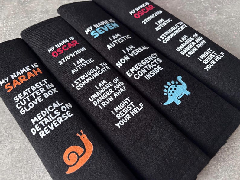May include: Four black rectangular seatbelt covers featuring embroidered text and graphics. Each cover displays a name, medical information, and safety warnings in white, orange, or blue thread. Designs include a snail and a dinosaur icon.