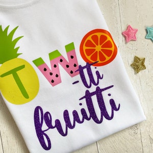 May include: White t-shirt with a purple glitter design that says "Two-tti fruiti" with a pineapple, watermelon, and orange graphic.