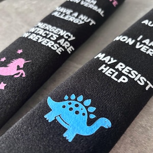May include: Two black fabric seatbelt covers featuring white text and colorful graphics. One cover displays a pink unicorn and text stating: I have a nut allergy, I am non verbal, emergency contacts are on reverse. The other shows a blue dinosaur.
