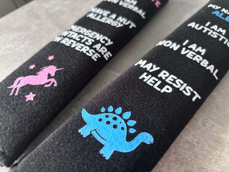 May include: Two black fabric seatbelt covers featuring white text and colorful graphics. One cover displays a pink unicorn and text stating: I have a nut allergy, I am non verbal, emergency contacts are on reverse. The other shows a blue dinosaur.