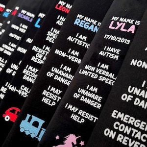 May include: A series of black fabric wristbands featuring white printed text that reads My Name Is followed by a name, I Am Autistic, Non-Verbal, I Am Unaware Of Danger, and Emergency Contact On Reverse. Some bands include colorful icons like trains or unicorns.