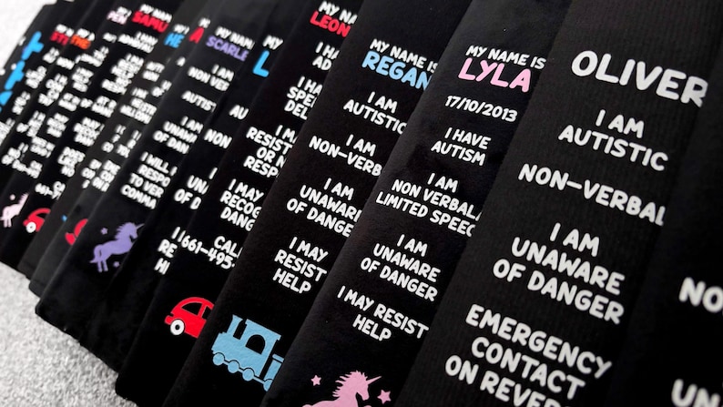 May include: A series of black fabric wristbands featuring white printed text that reads My Name Is followed by a name, I Am Autistic, Non-Verbal, I Am Unaware Of Danger, and Emergency Contact On Reverse. Some bands include colorful icons like trains or unicorns.